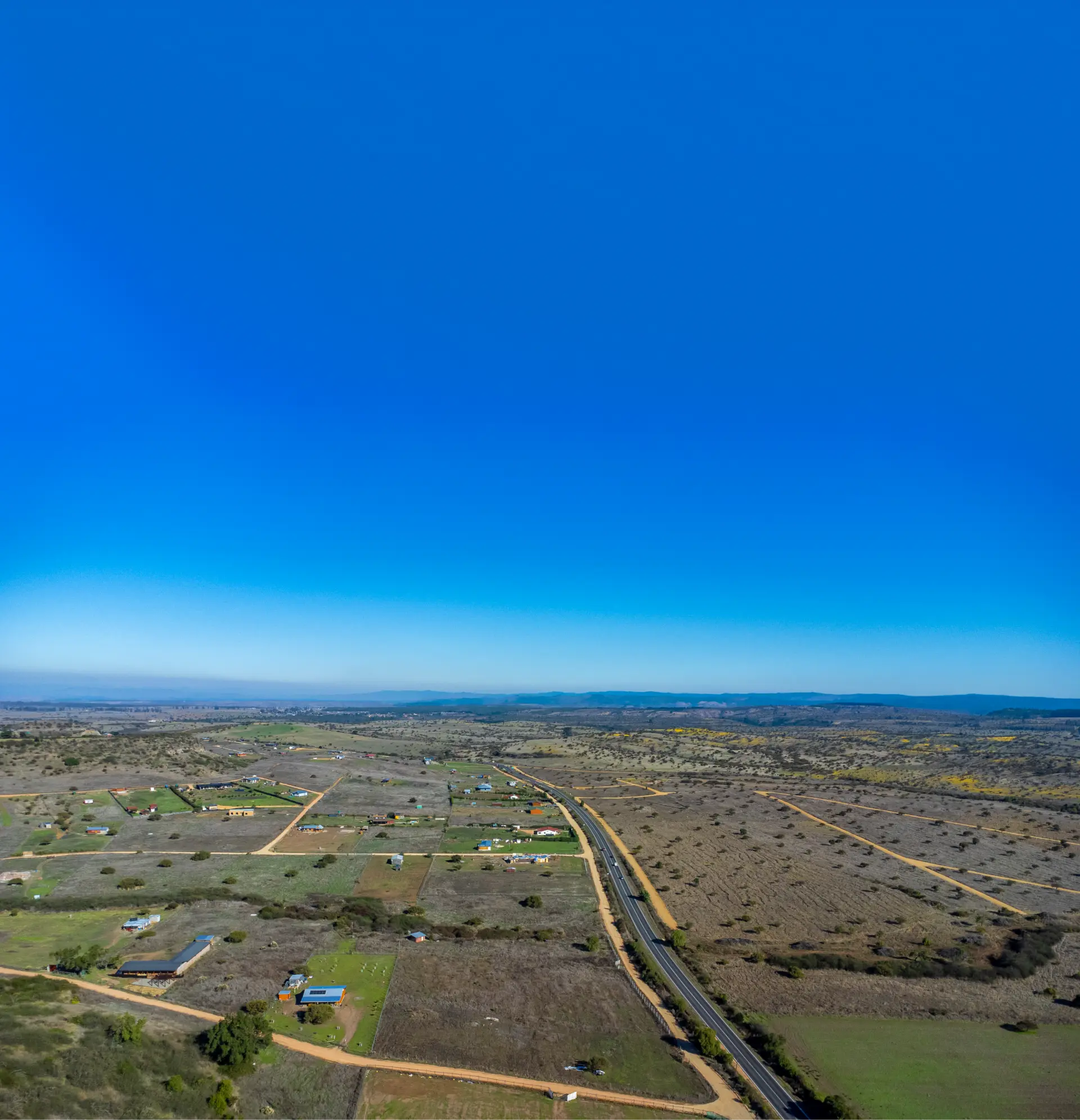 Vista aérea capturada con dron de parcelas planas y en lomaje en la Región de O'Higgins, ubicadas junto a la carretera, algunas con construcciones, bajo un cielo azul despejado.